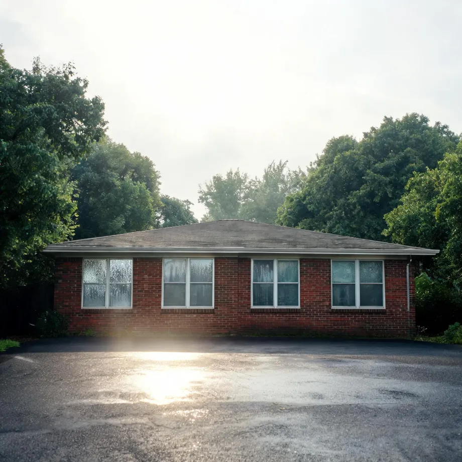 A single-story red brick building with a low hipped shingle roof and large windows, fronted by an empty asphalt parking lot with sun glare, surrounded by leafy green trees under a bright hazy sky.