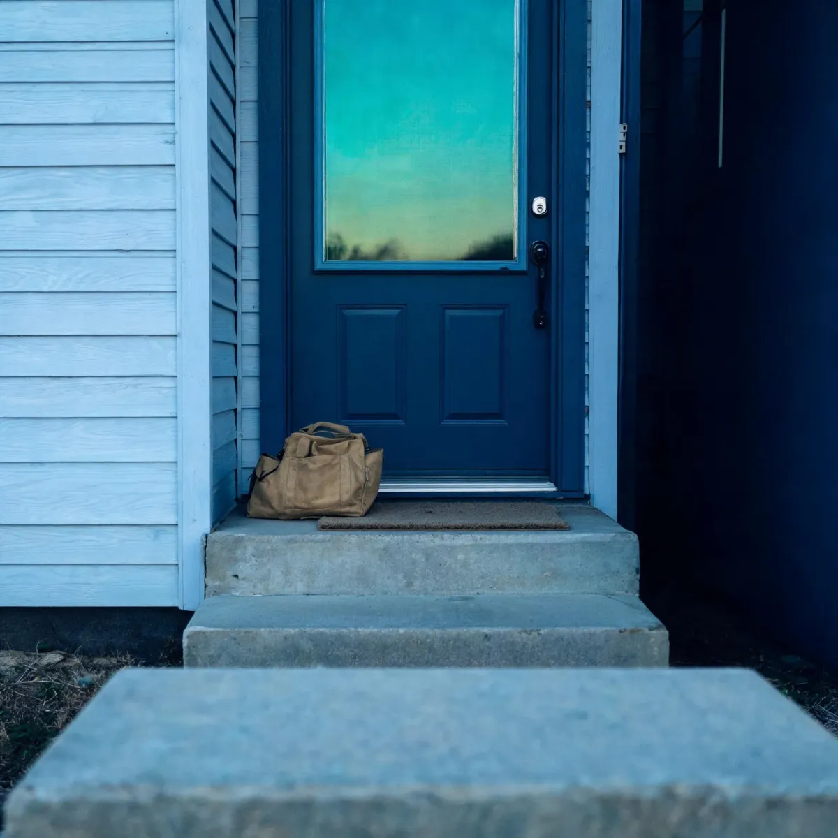 A tan duffel bag sits on a doormat in front of a closed dark blue door with a large window pane, beside pale horizontal siding and concrete steps.