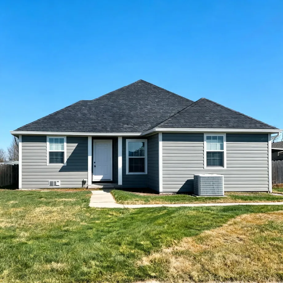 Single-story house with gray vinyl siding, dark shingled hip roof, white front door, two windows, an air conditioning unit beside the house, a concrete walkway, and a grass lawn under a clear blue sky.