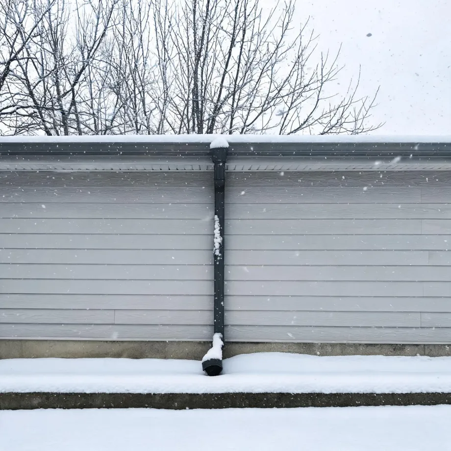 Exterior wall of a building with light gray horizontal siding and a dark downspout running to the ground, with snow on the roofline, steps, and ground, and bare trees behind during falling snow.