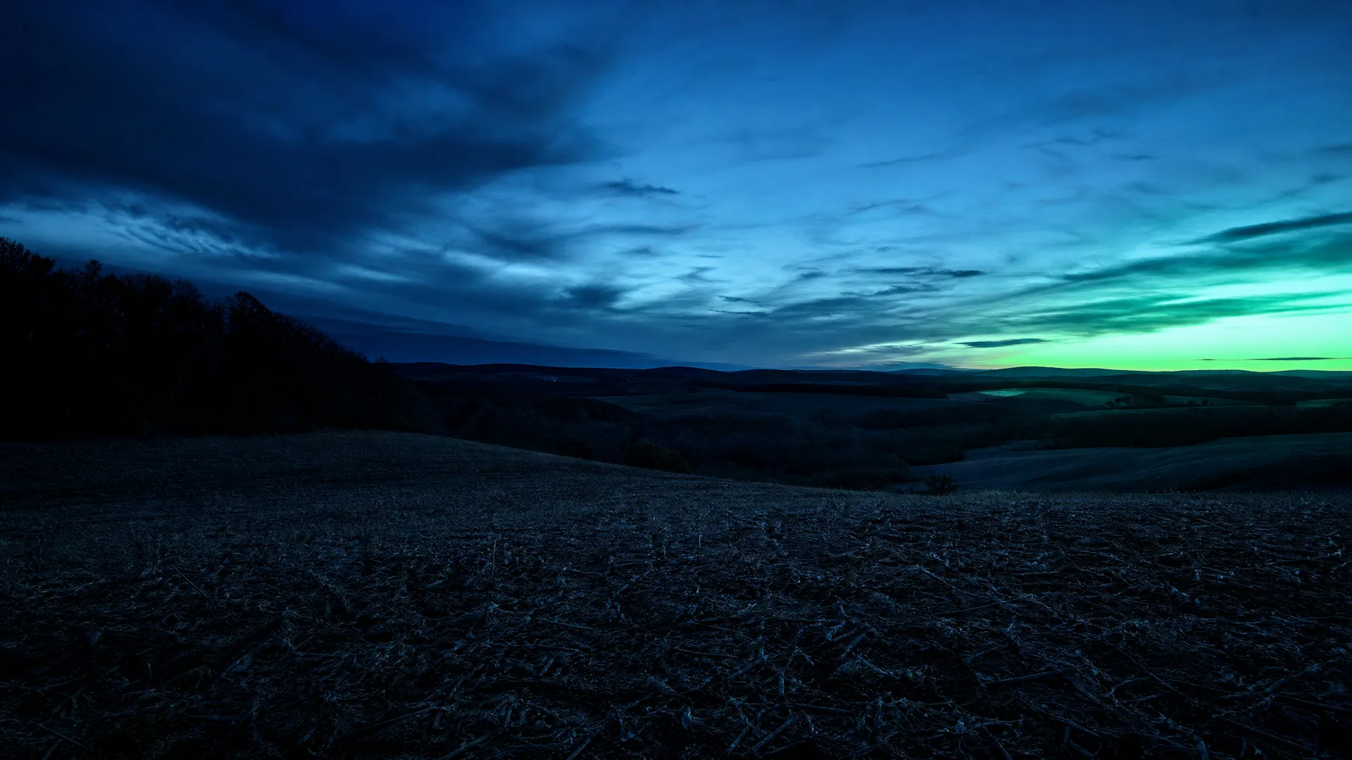A wide twilight landscape with a dark stubble field in the foreground, low rolling hills and trees beyond, under a cloudy blue sky with a green-yellow glow near the right horizon.