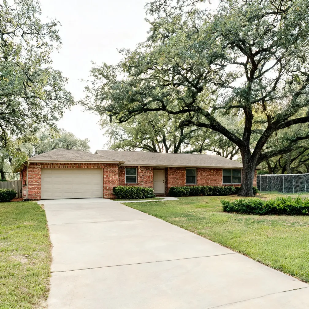 Single-story red brick house with a two-car garage, long concrete driveway, trimmed shrubs and a lawn, framed by large oak trees and a chain-link fence at right.