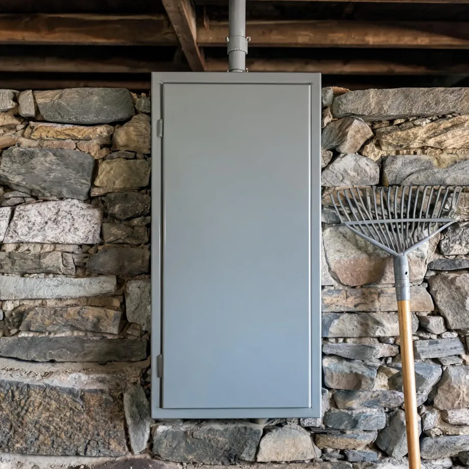 A gray metal cabinet with a vertical pipe rising from its top is mounted on a dry stone wall under wooden beams, with a leaf rake leaning against the wall to the right.