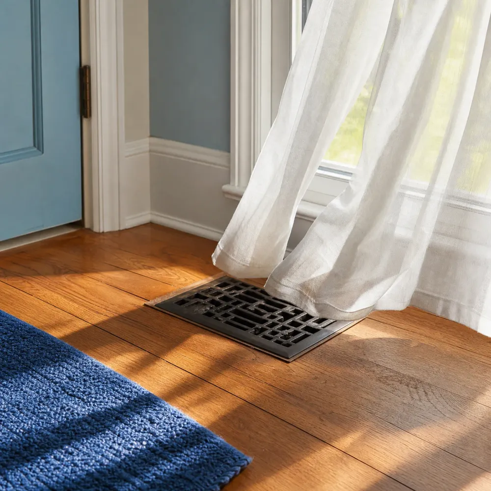 A dark metal floor register vent set into hardwood flooring near a window with sheer white curtains, with a blue rug in the foreground and a blue door at left.