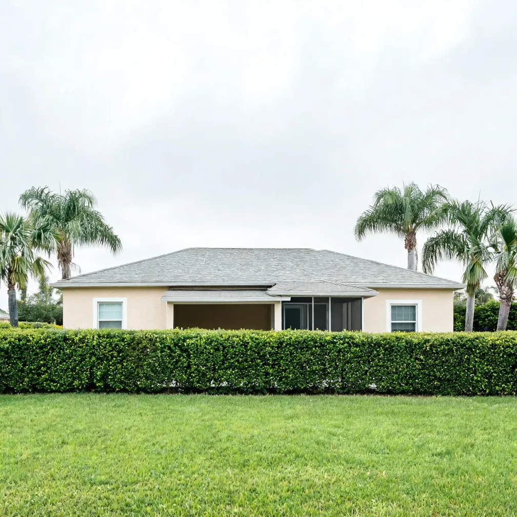 A single-story stucco house with a gray shingle hip roof, screened porch, and two windows, fronted by a trimmed hedge and mowed lawn, with palm trees on either side under an overcast sky.