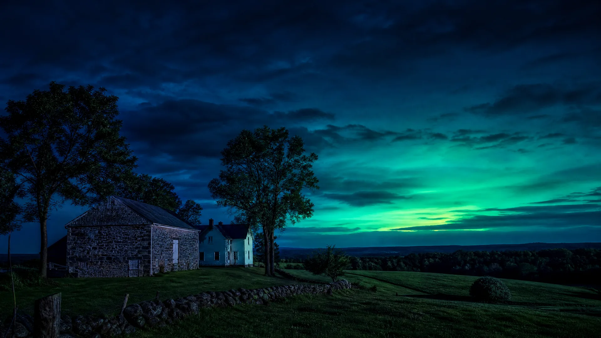 A stone barn and white farmhouse on a grassy hill at twilight, framed by bare-limbed trees, under a dark cloudy sky with a green glow near the horizon.