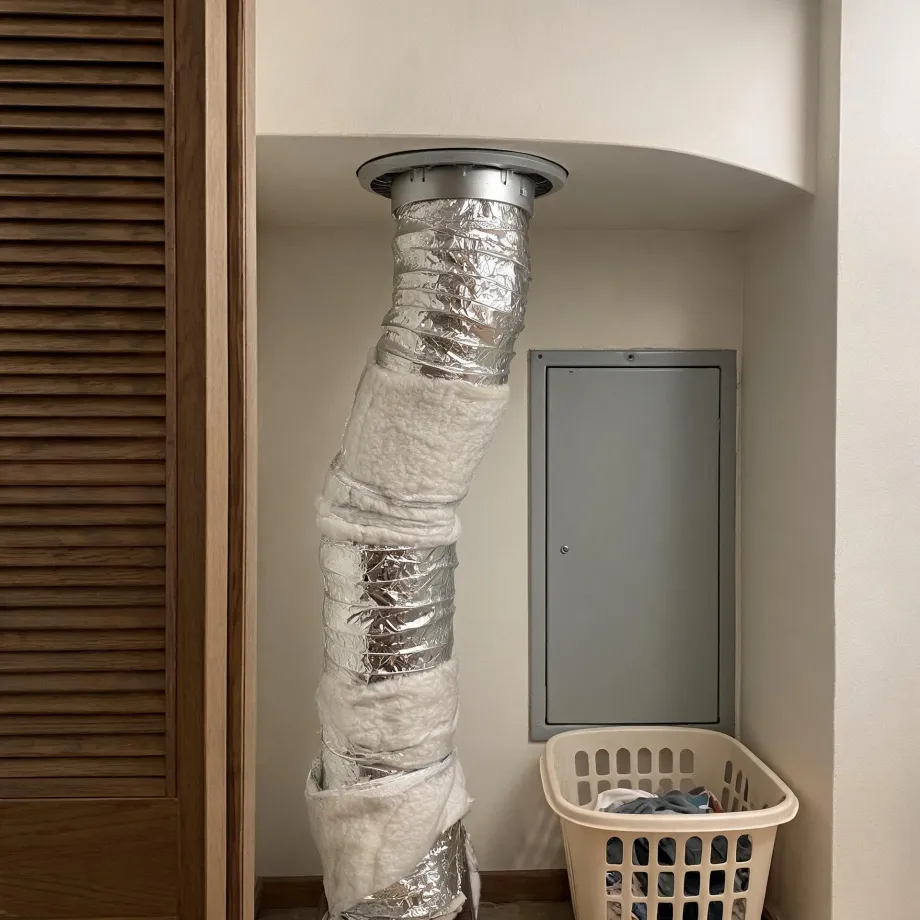 An interior alcove where a flexible foil duct runs from a round ceiling collar down to the floor, partly wrapped in white insulation, next to a gray metal wall access panel, a louvered wooden door, and a beige laundry basket holding clothes.