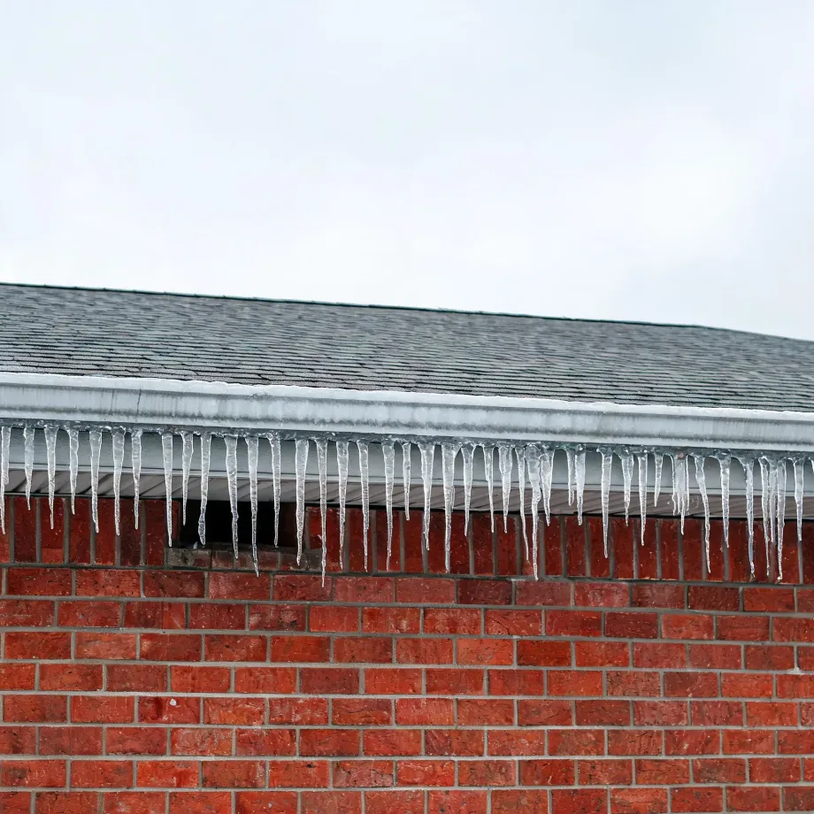 A row of icicles hangs from the white gutter and soffit along the edge of an asphalt-shingle roof above a red brick wall, under an overcast white sky.
