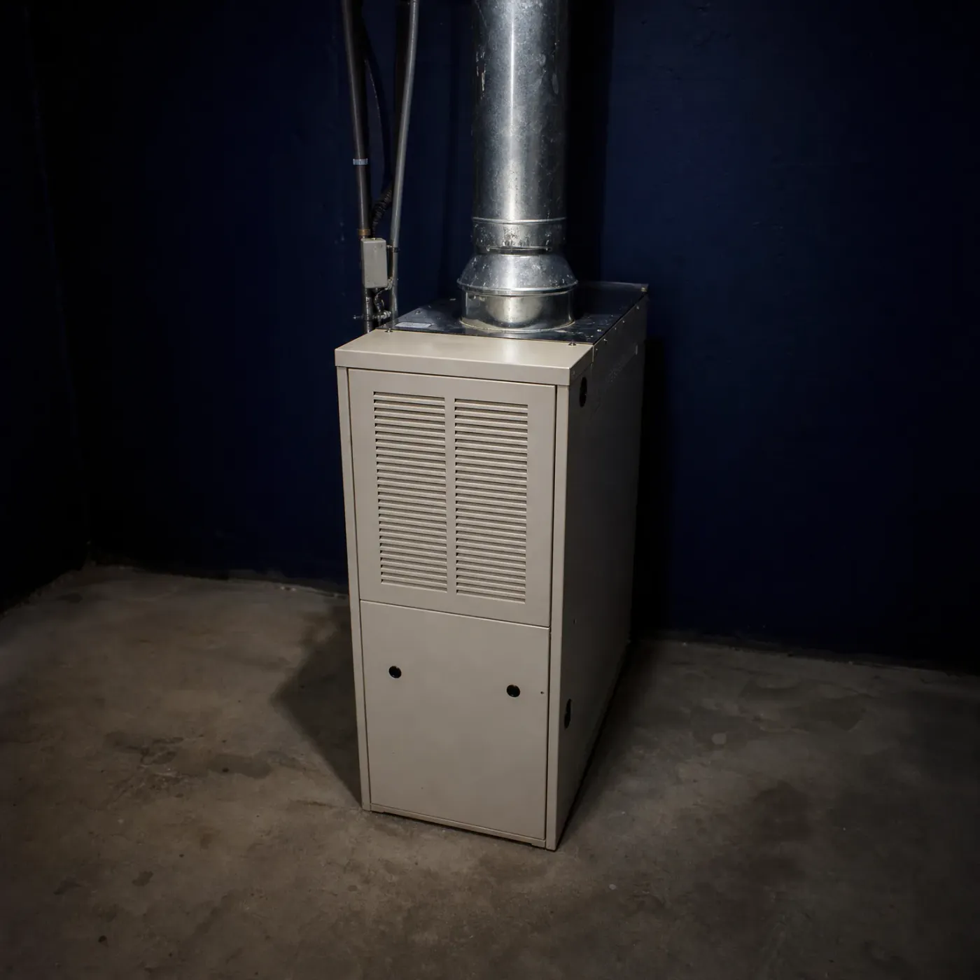 A beige residential furnace with a louvered front panel stands on a concrete floor in a basement corner with dark blue walls, connected to a galvanized metal flue pipe rising to the ceiling.