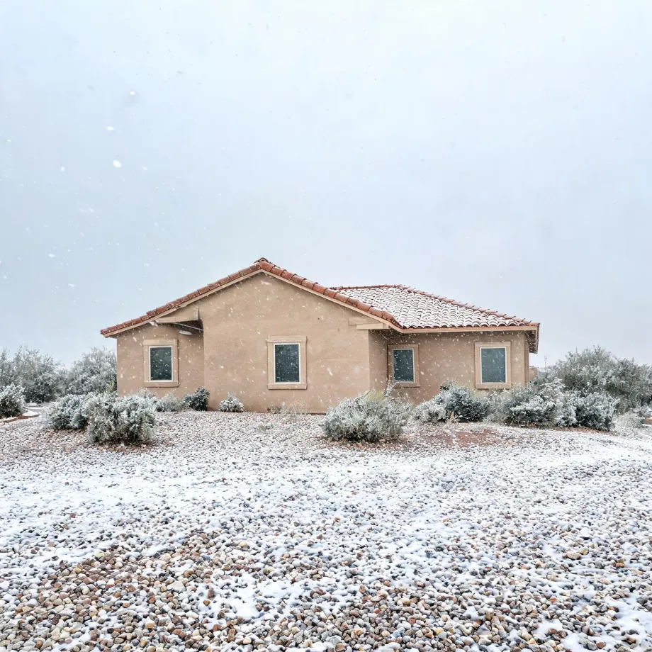 A single-story tan stucco house with a red tile roof stands behind snow-dusted desert shrubs and a gravel yard during falling snow under an overcast sky.