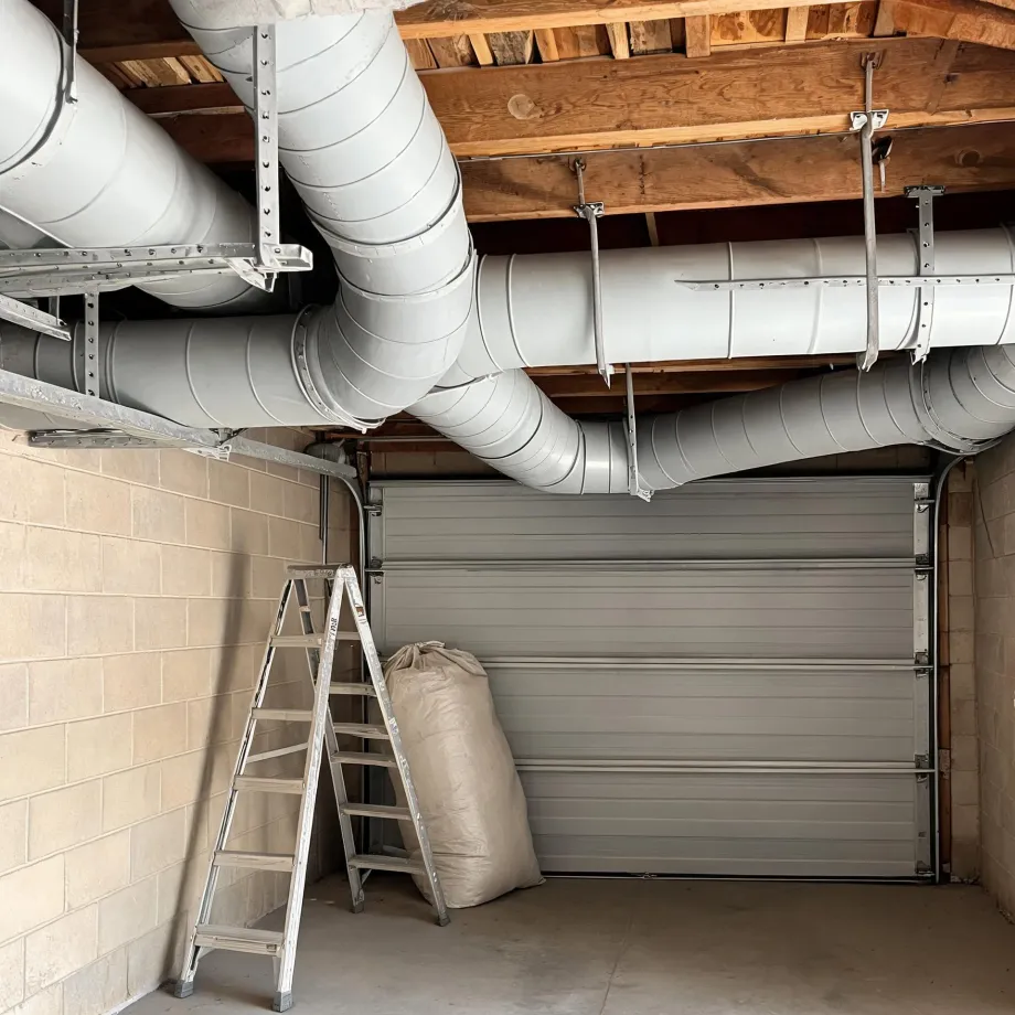 Interior of a garage with large round metal ducts suspended from a wooden joist ceiling, a concrete block wall, a sectional garage door, an aluminum step ladder, and a filled fabric sack on the floor.