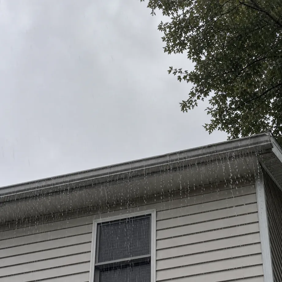 Upward view of the gable end and gutter line of a house with light horizontal vinyl siding, with water streaming off the overflowing gutter against an overcast sky and tree branches at the upper right.