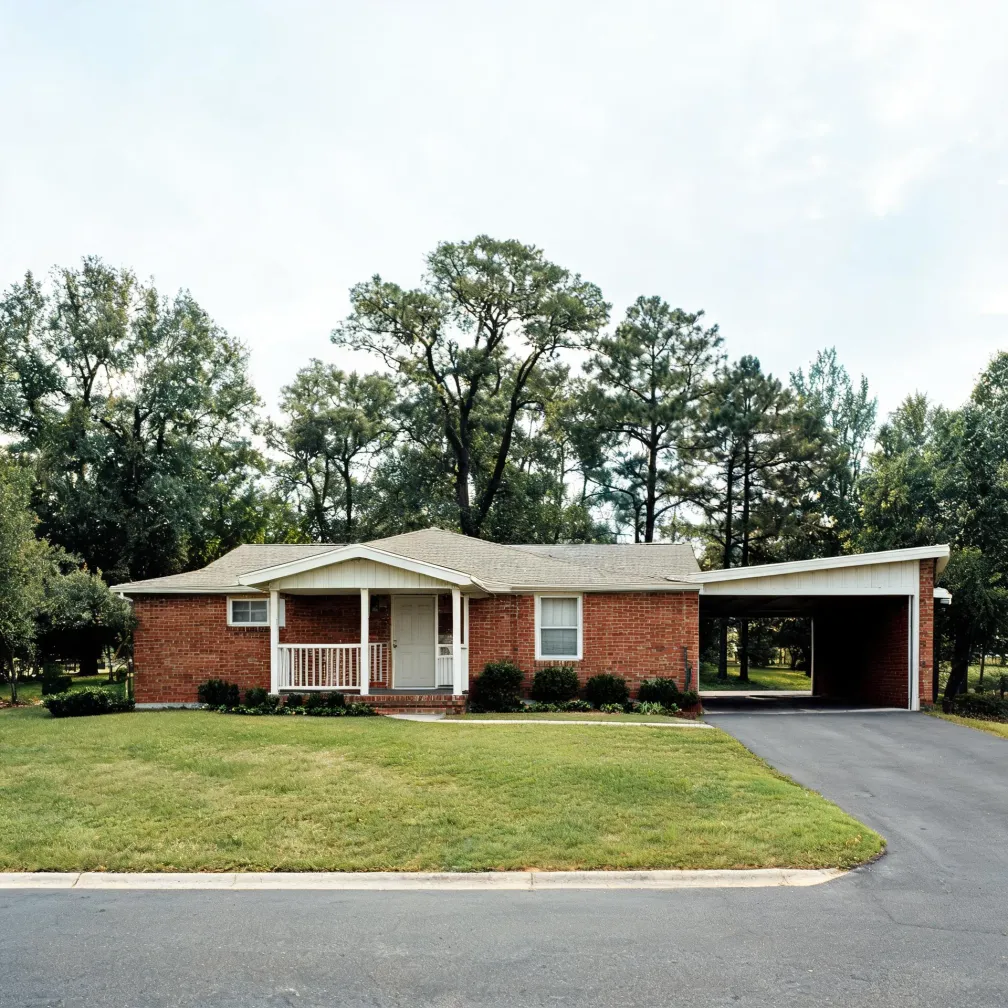 A single-story red brick ranch house with a small white-columned front porch and an attached carport, set behind a mown lawn with an asphalt driveway and street in front and tall deciduous and pine trees behind.