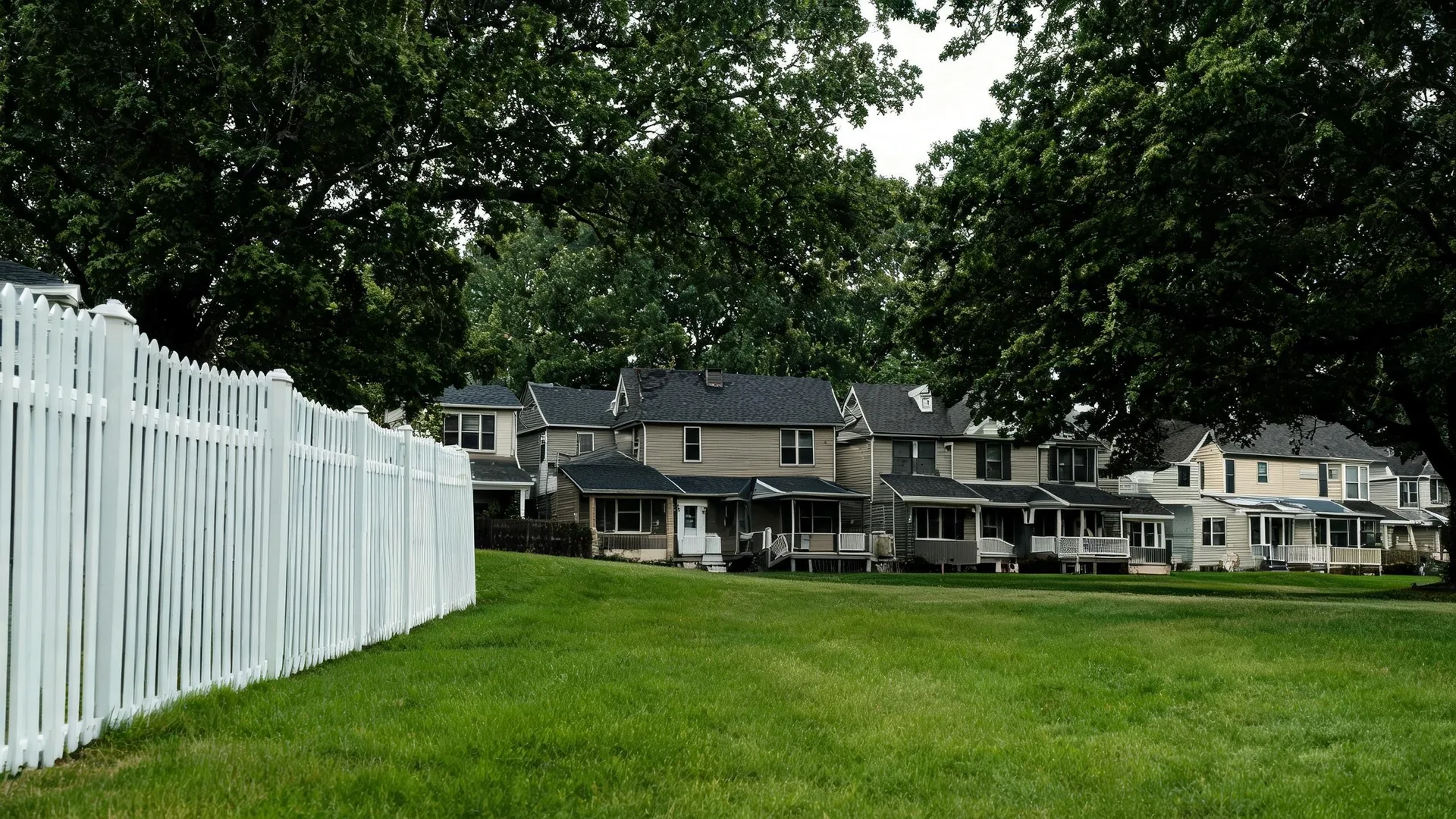 A white picket fence runs along the left edge of a mowed grass lawn, with a row of two-story houses with porches and large leafy trees behind them.