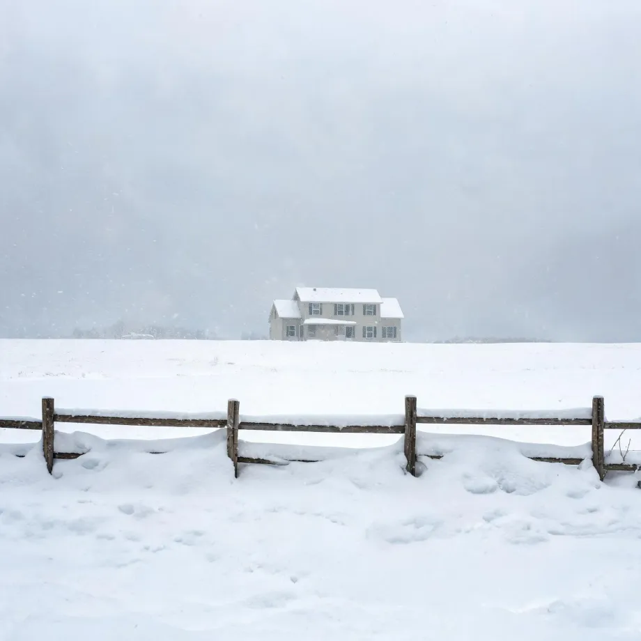 A two-story light-colored house with a snow-covered roof stands alone on a flat snow-covered field under a pale gray sky, with a weathered wooden post-and-rail fence partly buried in snow in the foreground.