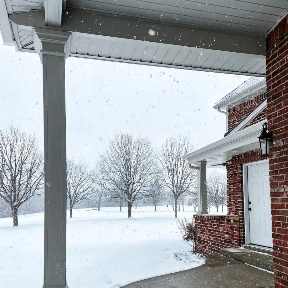 View from under a covered porch with a gray column toward a snow-covered lawn with bare trees, with a red brick house wall, white door, and wall lantern at right during falling snow.