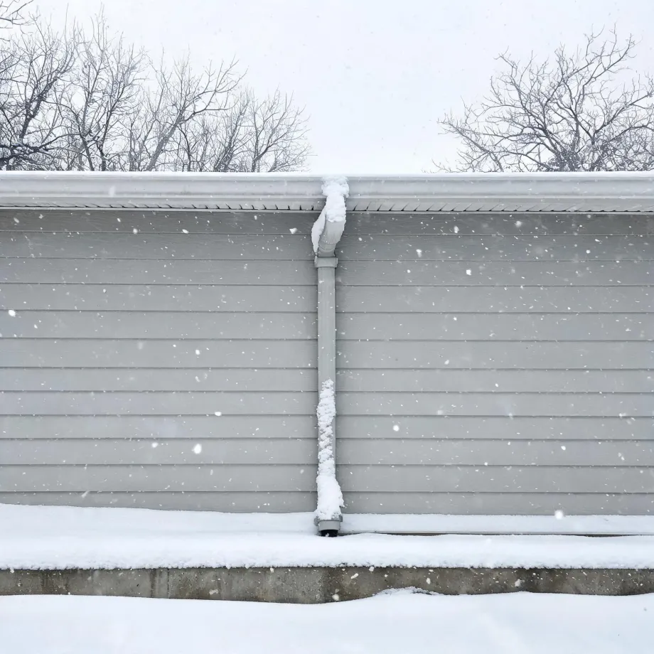 Exterior wall of a building with light gray horizontal lap siding and a downspout running from the snow-covered gutter to the ground, with bare trees behind the roofline and snow falling and accumulating on the ground.