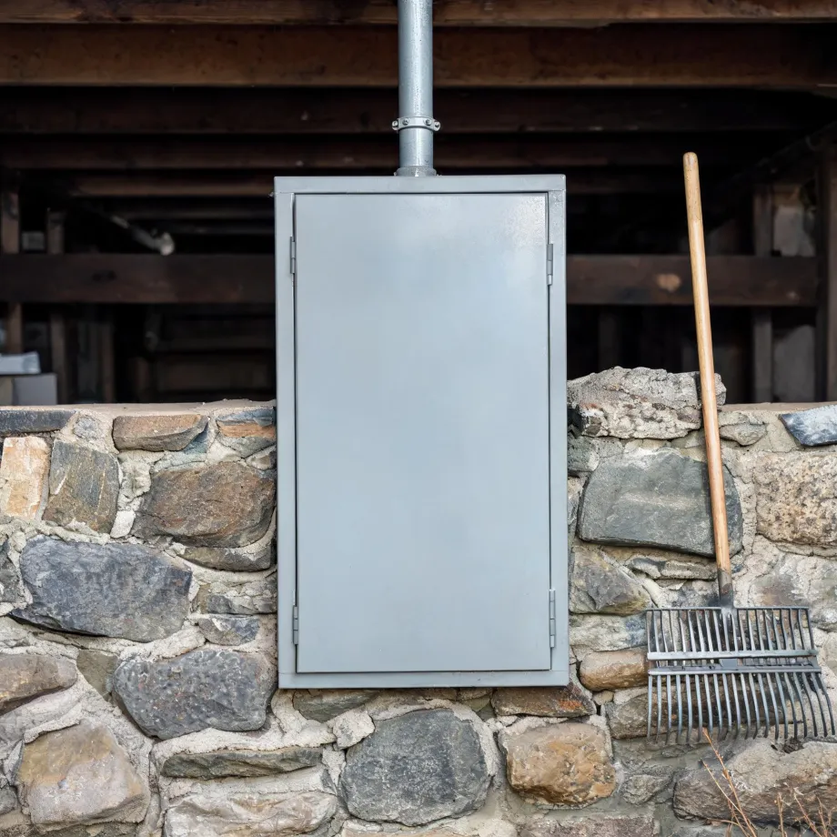 A gray metal utility cabinet with a vertical pipe rising from its top is mounted on a stone wall, with a rake leaning against the wall to the right and dark wooden beams in the shaded space behind.