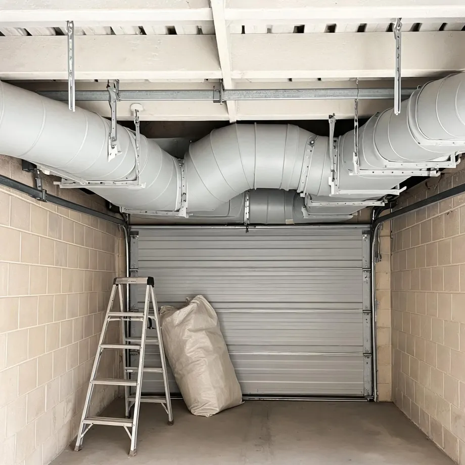 Interior of a garage with concrete block walls, a closed metal roller door, large round ventilation ducts suspended from the ceiling, an aluminium stepladder and a filled white sack on the floor.
