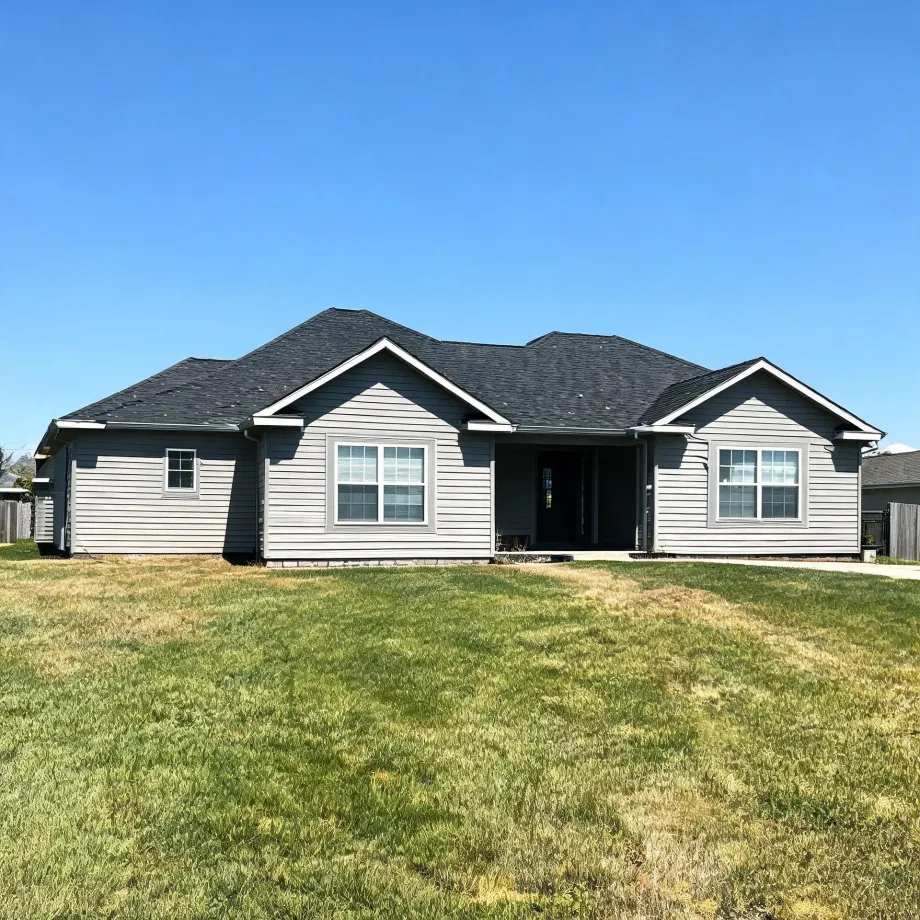 Single-story gray horizontal-siding house with dark shingled gable roof, white-trimmed windows and a recessed front entry, fronted by a large green lawn under a clear blue sky.