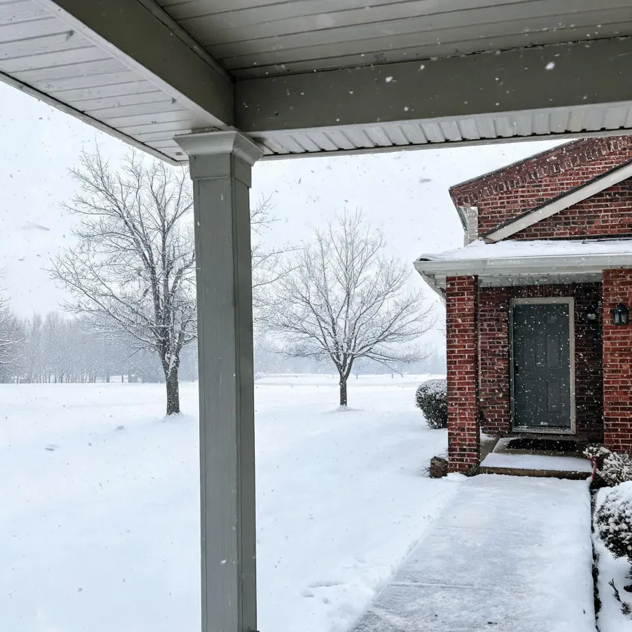 View from under a covered porch with a white column toward a red brick house with a dark door, a snow-covered lawn, walkway, and bare trees during falling snow.