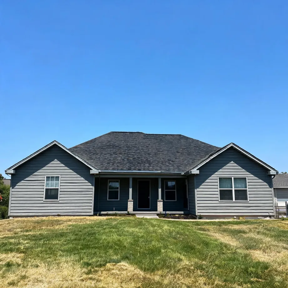 A single-story gray lap-sided house with a dark shingled hip roof, two front-facing gables, and a small covered porch with white columns, set behind a patchy green and dry lawn under a clear blue sky.