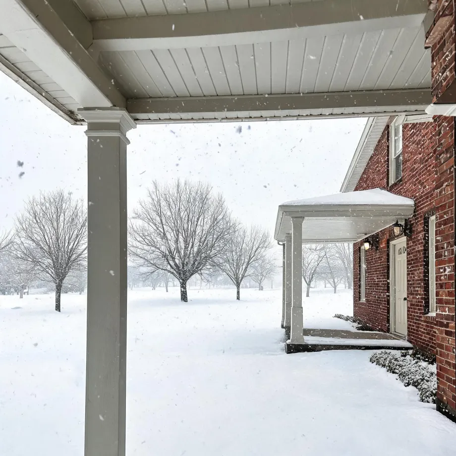 View from under a covered porch with a white column toward a red brick house with a white entry door and portico, with snow falling over a snow-covered lawn and bare trees.