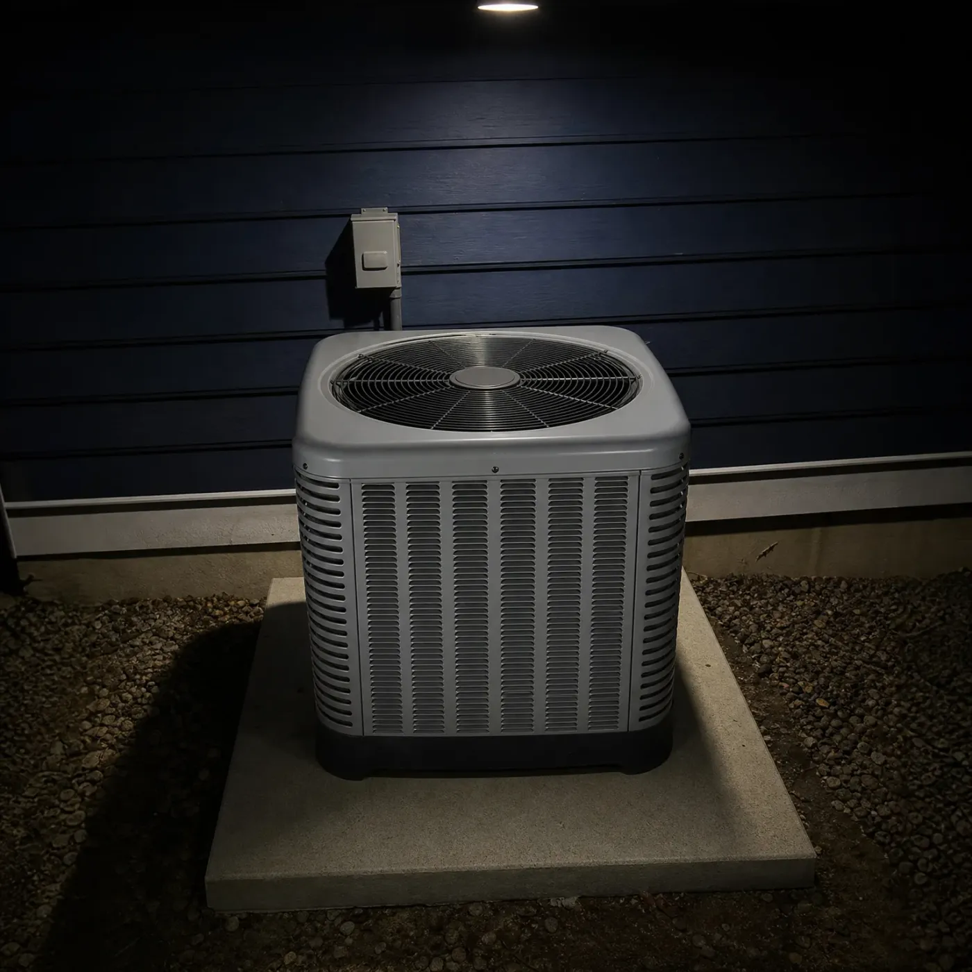 A gray outdoor condensing unit with a top-mounted fan grille sits on a concrete pad surrounded by gravel, against dark horizontal siding with a wall-mounted electrical disconnect box above it, lit at night.