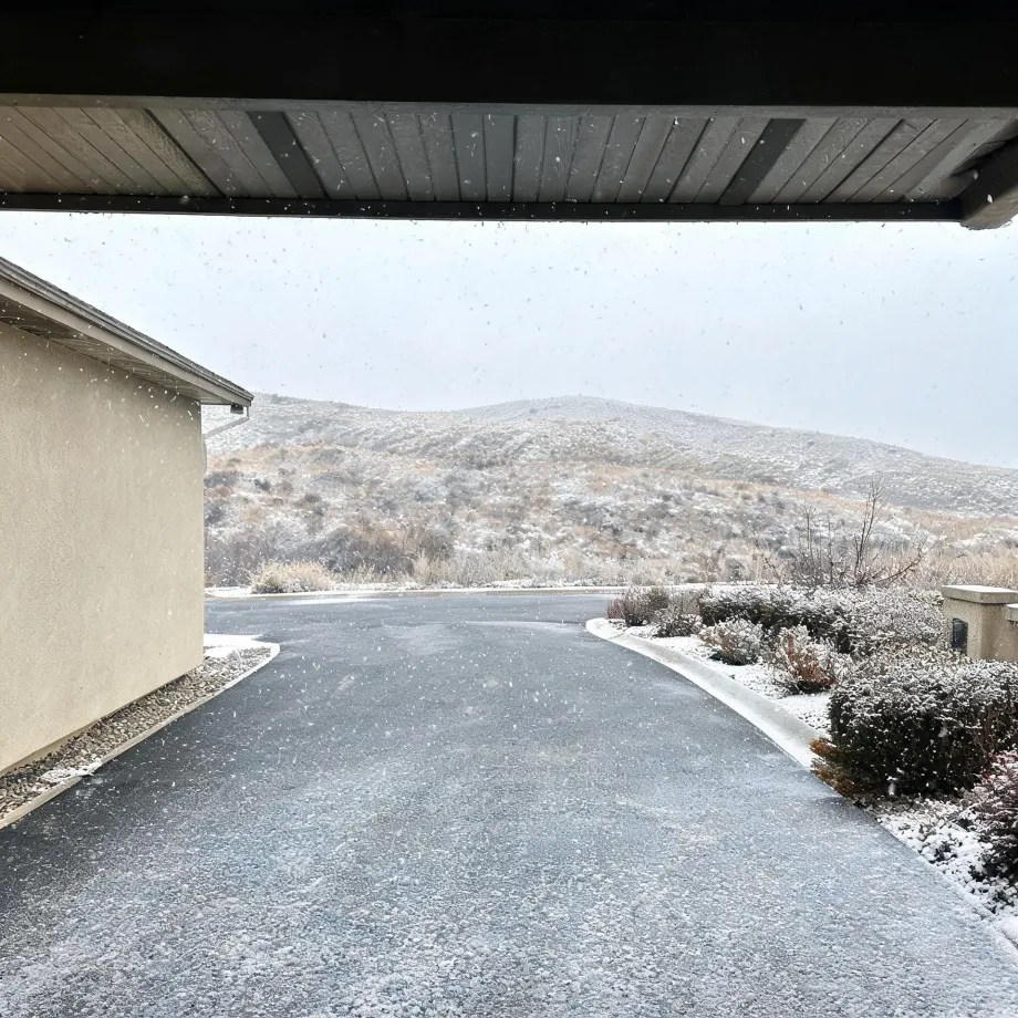 View from beneath a covered entry of a paved driveway dusted with snow, with a stucco wall at left, snow-covered shrubs at right, and low scrub-covered hills under an overcast sky with falling snowflakes.