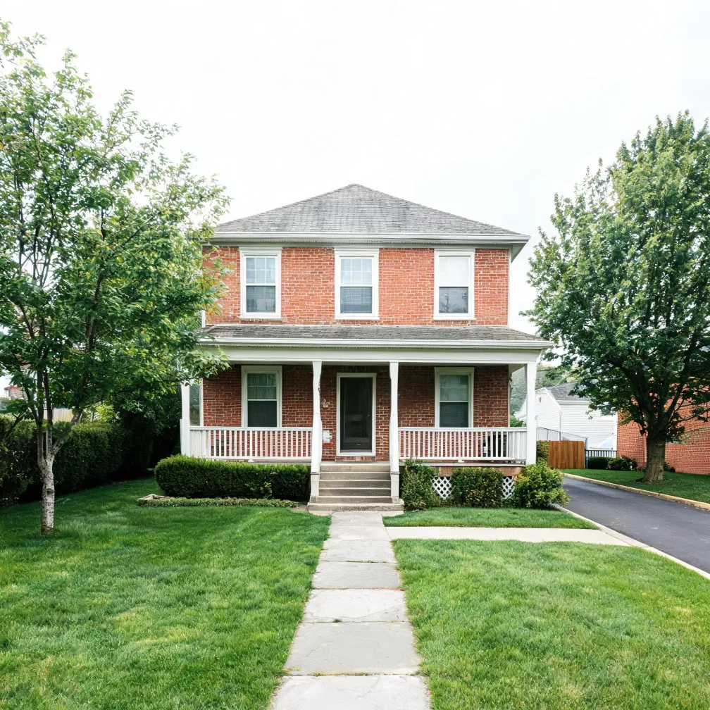 A two-story red brick house with a white covered front porch, gray hipped roof, and a stone walkway leading across a mown green lawn, with deciduous trees on both sides and a driveway at right.