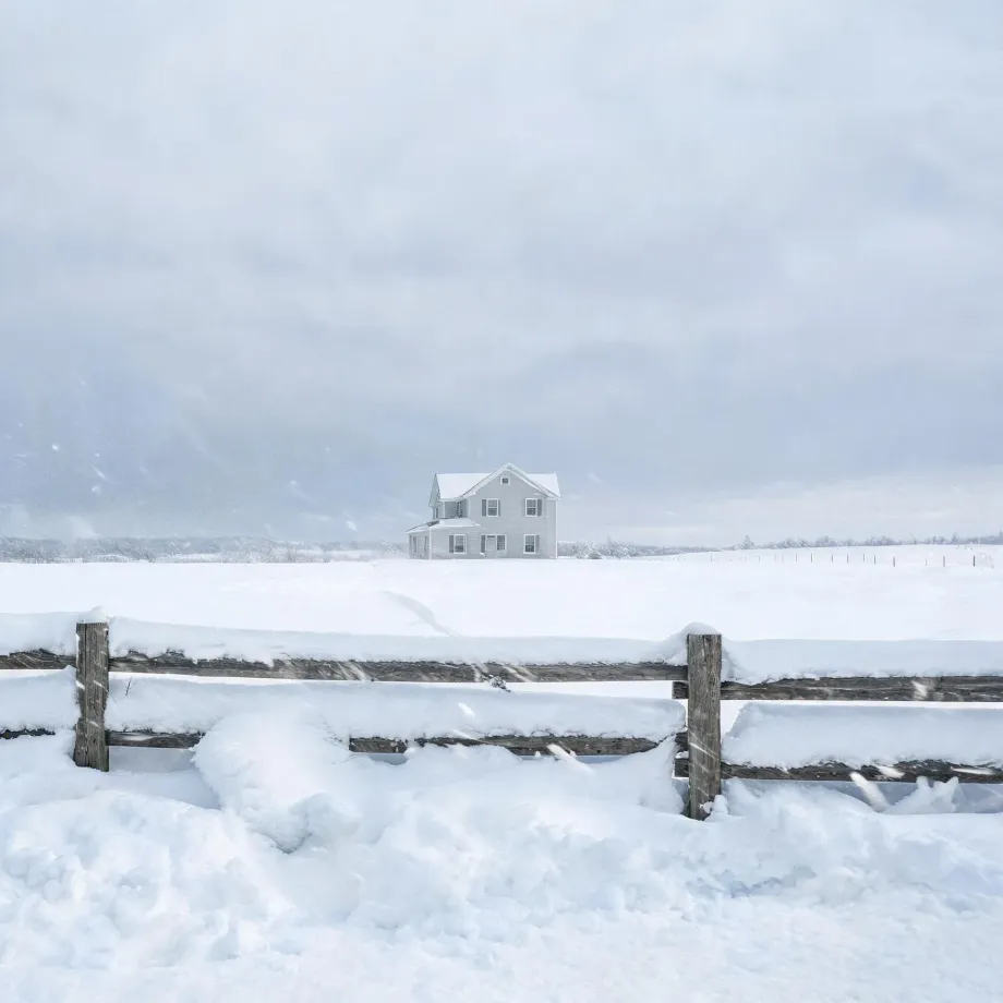 A two-story light-colored house stands alone in a snow-covered field under an overcast sky, with a snow-topped wooden rail fence in the foreground.