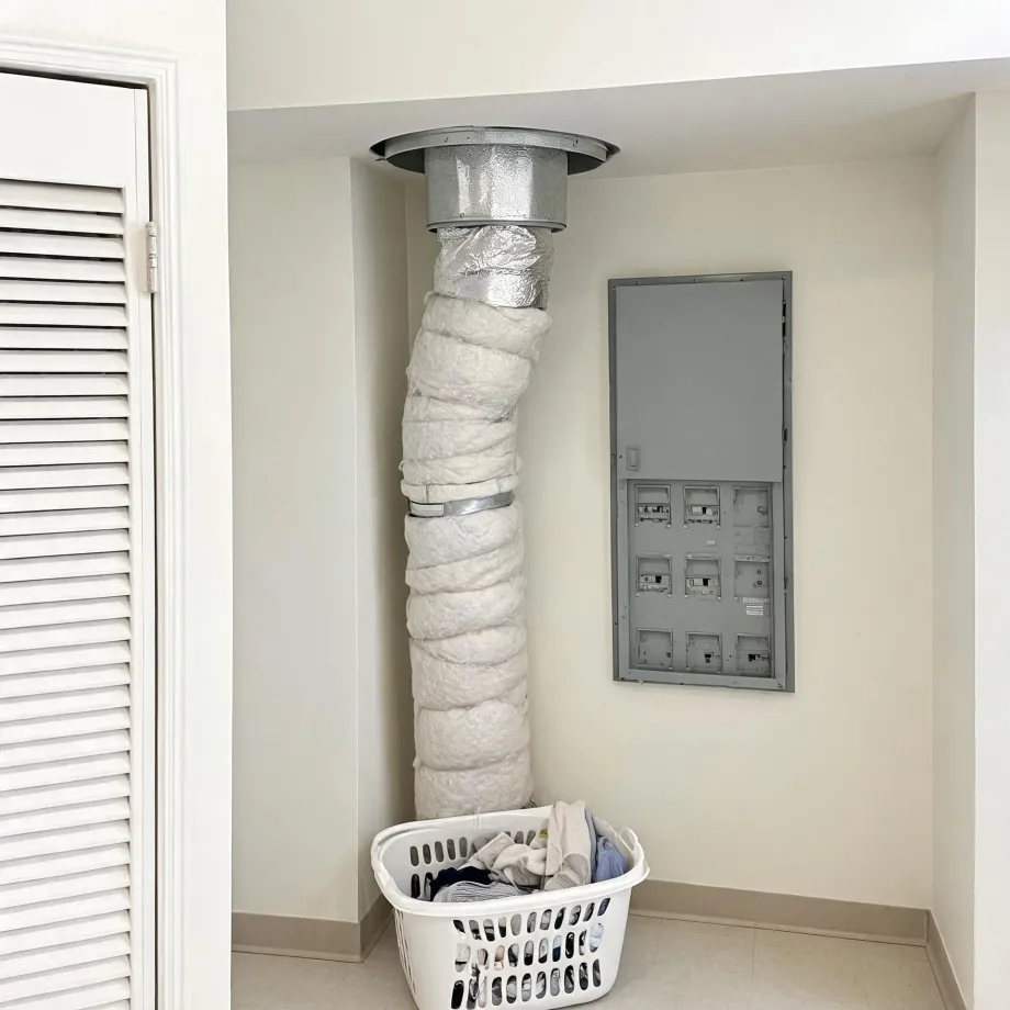 An interior corner of a room where an insulation-wrapped duct runs from the floor area up to a metal collar in the ceiling, beside a gray electrical panel on the wall, with a white laundry basket of clothes on the tile floor and a louvered door at left.