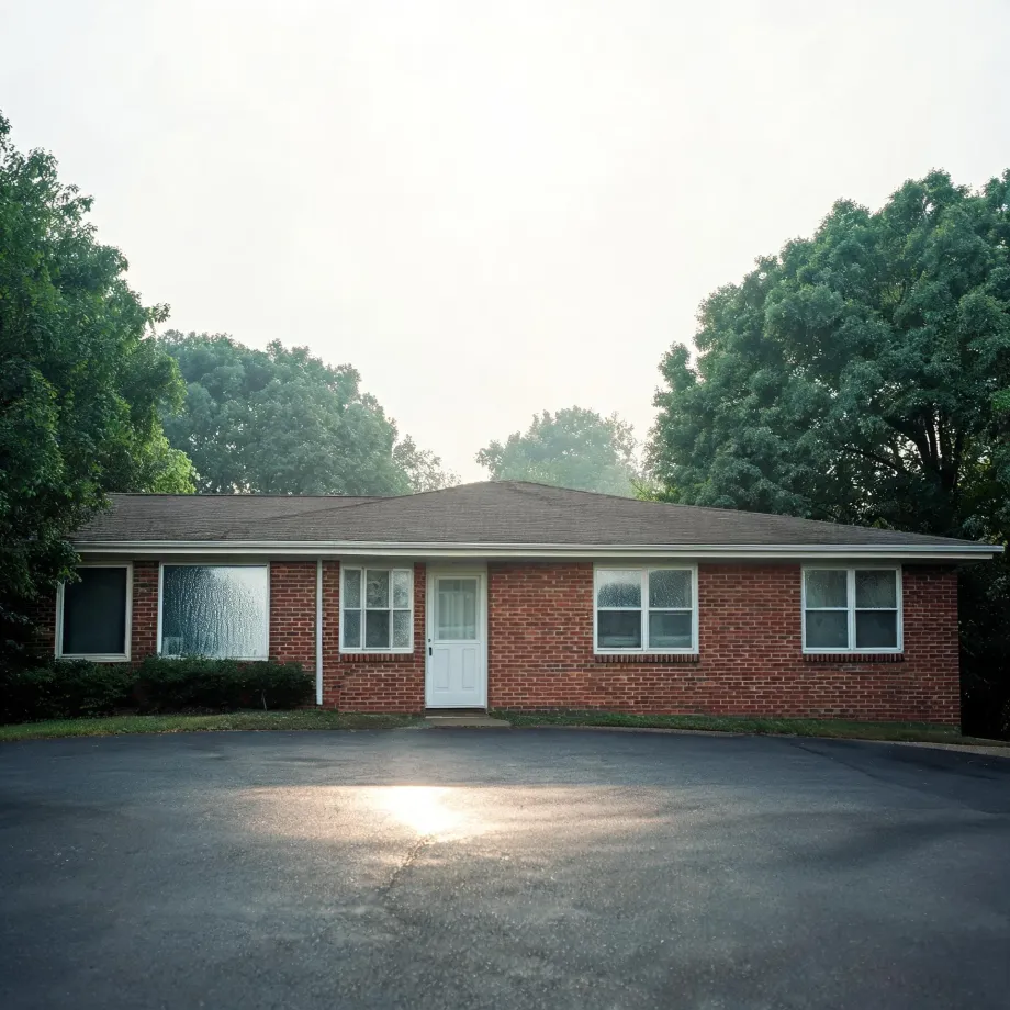 A single-story red brick ranch house with a white front door, white-framed windows and a shallow shingled roof, fronted by a wide asphalt parking area and backed by leafy green trees under a bright hazy sky.