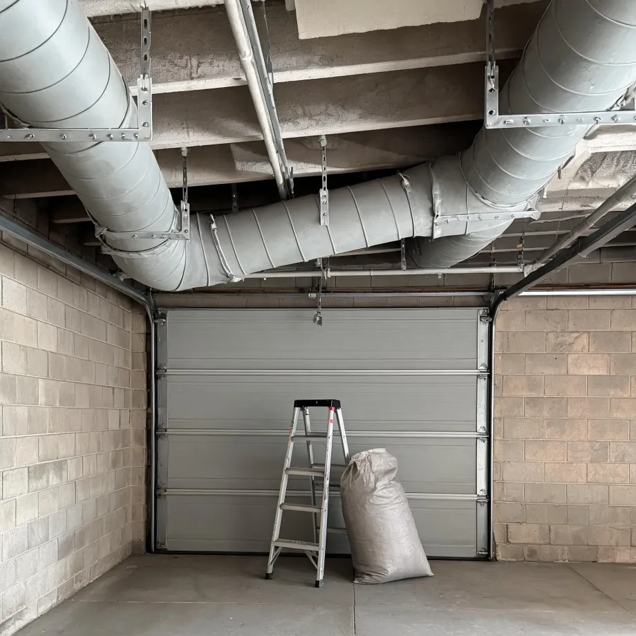 Interior of a concrete block garage with a closed sectional door, a folding step ladder and a filled burlap sack on the floor, and large metal ductwork suspended from the ceiling.