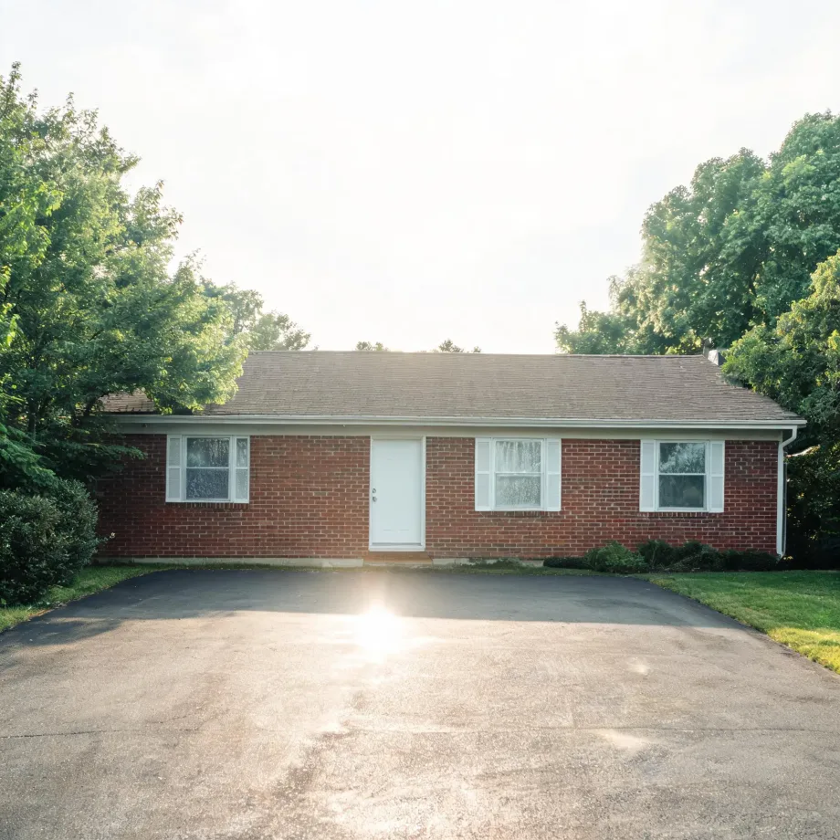 A single-story red brick ranch house with a white front door, white-trimmed windows and shingled roof, fronted by a wide asphalt driveway with sun flare, flanked by green trees and grass.