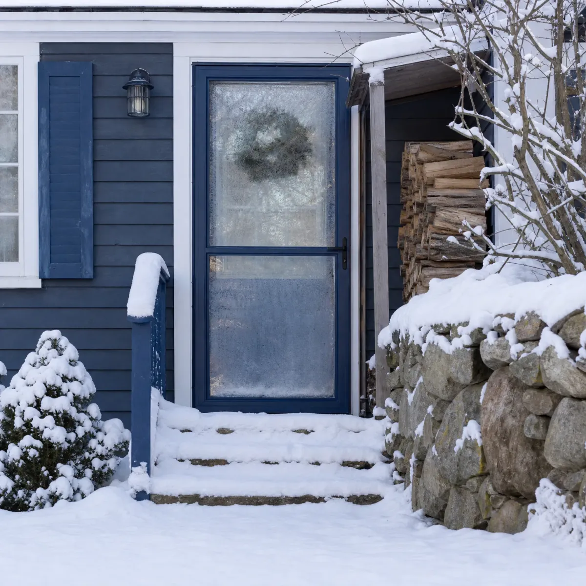 Snow-covered stone steps lead to a blue storm door on a blue clapboard house, with a stacked woodpile, a snowy stone wall, and bare shrubs nearby.