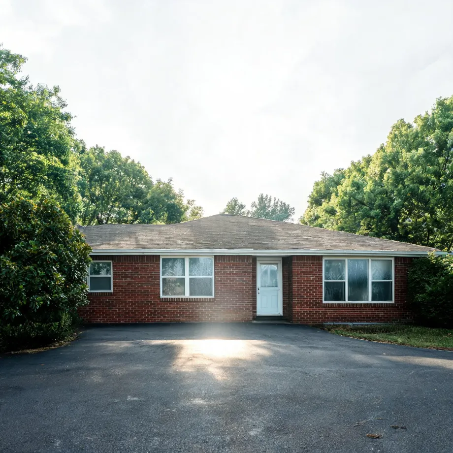 A single-story red brick house with a hipped shingle roof, white-framed windows and a pale blue front door, fronted by a wide asphalt driveway and flanked by green trees and shrubs under a bright hazy sky.