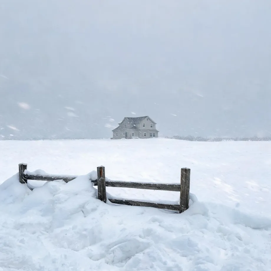 A two-story gabled house stands alone across a snow-covered field during falling snow, with a weathered wooden post-and-rail fence partly buried in a snowdrift in the foreground.
