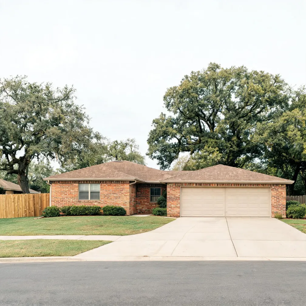 Single-story brick house with a shingled hip roof, two-car garage, wide concrete driveway, mown lawn, wooden fence at left, and large oak trees behind under an overcast sky.