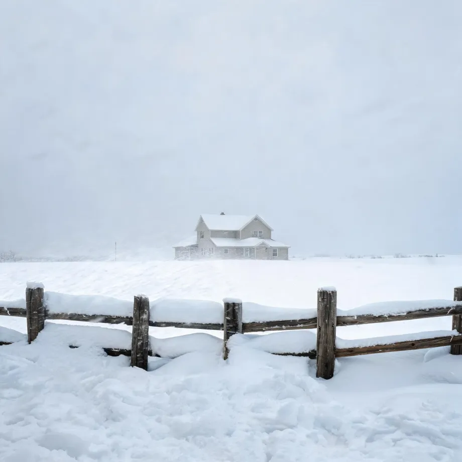 A two-story house stands in a snow-covered field under a pale hazy sky, with a snow-topped wooden rail fence crossing the foreground.