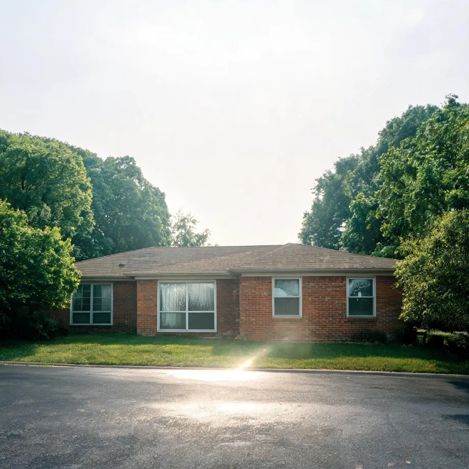 A single-story brick house with a shingled roof and white-framed windows, set behind a mown lawn and an asphalt street, flanked by large deciduous trees under a bright hazy sky with sun flare.