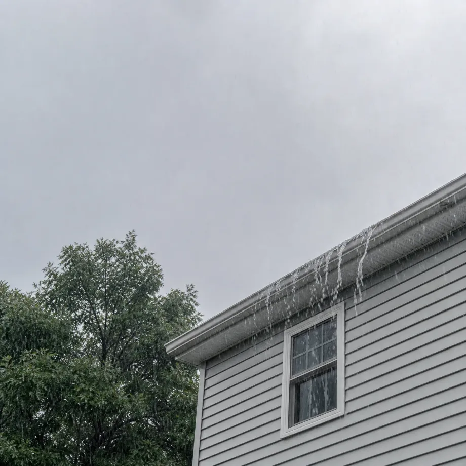 Water overflows from the gutter along the roof edge of a light gray vinyl-sided house with a white-framed window, under an overcast sky, with a leafy tree at left.