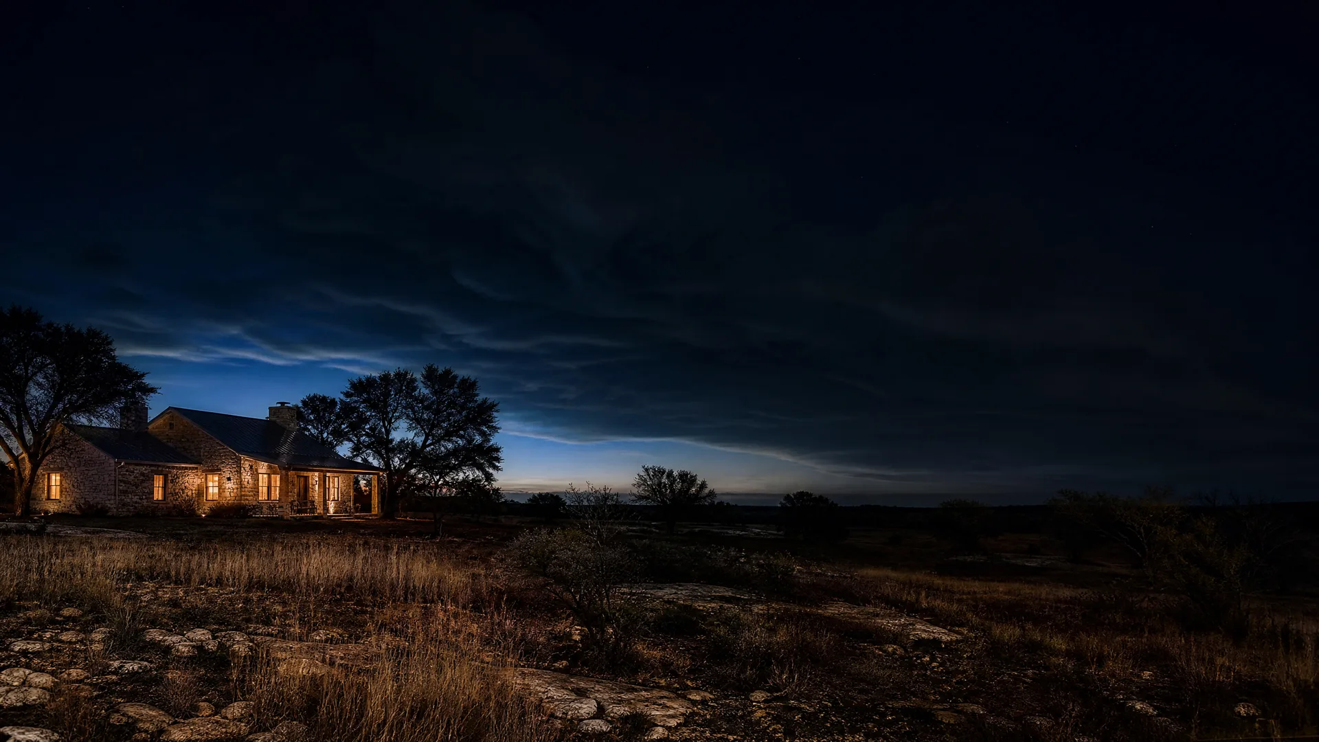 A single-story stone house with warmly lit windows stands among bare trees at twilight, with dry grass and rocky ground in the foreground and a band of blue light along the horizon beneath dark clouds.