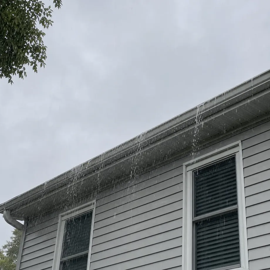 Water overflowing from the gutter along the eave of a light gray vinyl-sided house with two white-framed windows, seen from below against an overcast sky with tree branches at upper left.
