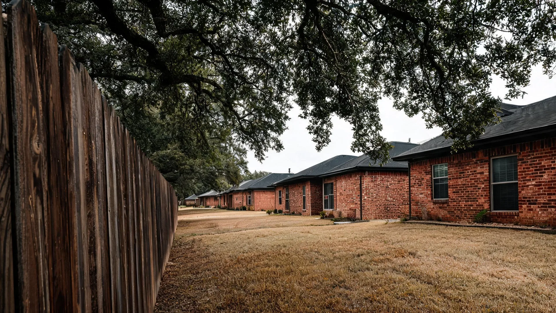 A row of single-story red brick buildings with dark shingled roofs stands along a dry brown lawn beside a weathered wooden fence, under overhanging oak branches and an overcast sky.