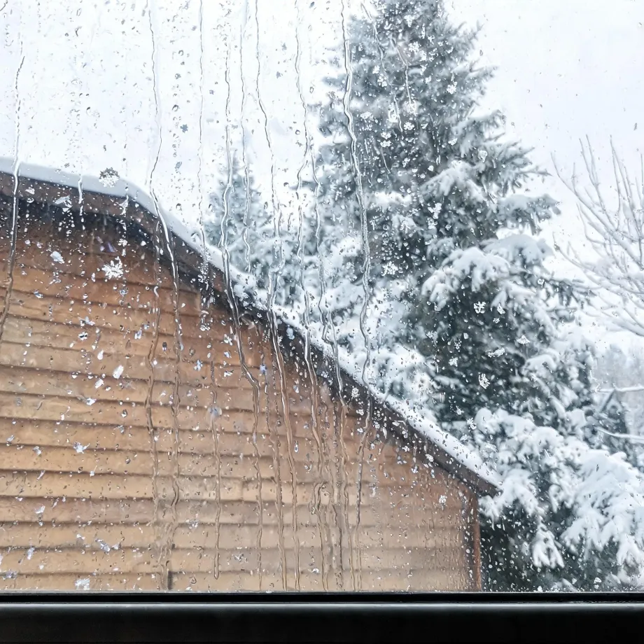 View through a wet, streaked window pane of a wooden clapboard building with a snow-covered roof and snow-laden conifer trees under an overcast sky.