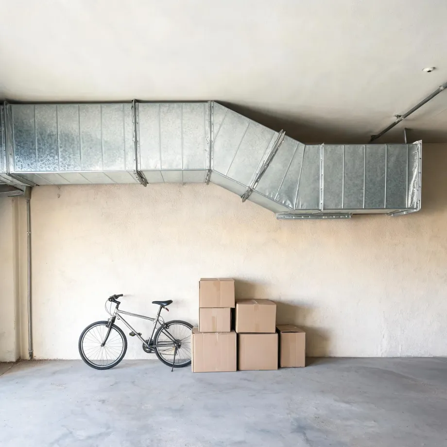 Interior of a garage with a large galvanized metal duct running along the ceiling, a bicycle leaning against a beige stucco wall, and six stacked cardboard boxes on a concrete floor.