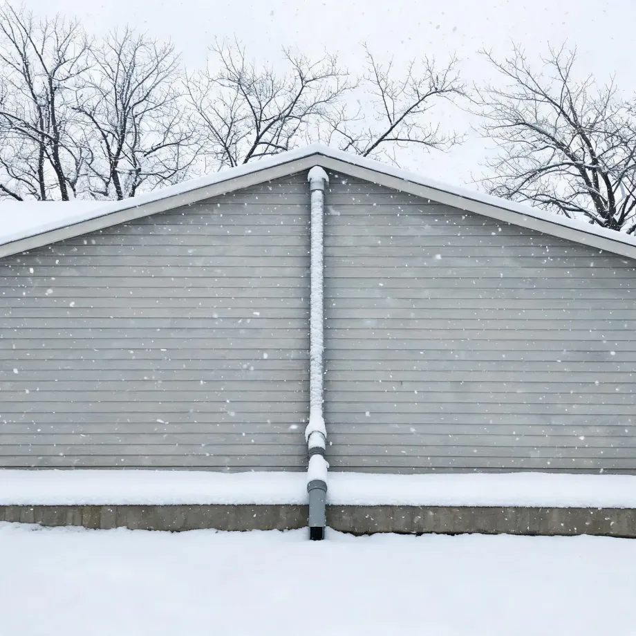 Gable end of a gray horizontal-sided building with a vertical downspout on its center, snow on the roof and ground, and bare trees behind during falling snow.