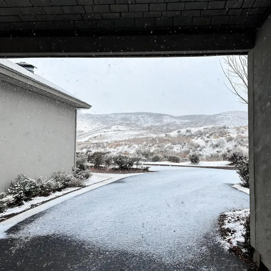 View from under a covered entry or carport toward a snow-dusted driveway, a stucco-clad building wing on the left, low shrubs, and snow-covered hills under a pale sky with falling snowflakes.