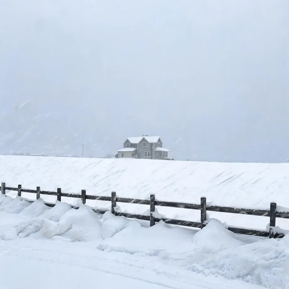 A two-storey house with a gabled roof stands on a snow-covered slope behind a dark wooden post-and-rail fence, under a pale hazy sky during snowfall.